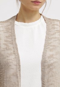 Beige knitted cardigan with an open front, featuring a textured pattern, layered over a white, textured fabric top.