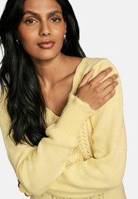 Maglione giallo a maglia con scollo a V, dotato di un motivo testurizzato e maniche ampie. Il modello ha lunghi capelli scuri e trucco naturale.