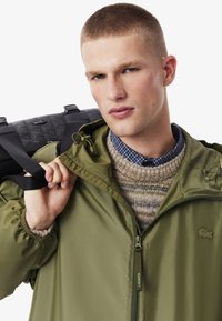 Giacca leggera verde oliva con polsini elasticizzati, zip frontale e logo Lacoste. Indossata sopra un maglione lavorato a maglia beige a motivi e una camicia a scacchi.