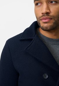 Mörkblå texturerad kappa med spetsig krage, som har en knappstängning och bärs över en grå tröja.