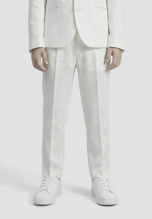 Man draagt witte pakbroeken, een bijpassend wit colbert en witte vetersneakers, staand op een effen grijze achtergrond.