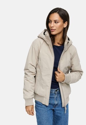 Veste à capuche beige avec une fermeture éclair à l'avant, des poignets côtelés et deux poches latérales. Portée par-dessus un haut bleu marine et associée à un jean bleu.