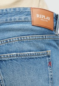 Jean en denim bleu clair avec une étiquette en cuir marron portant la marque "REPLAY" et des détails de couture. Poche arrière avec un accent logo rouge.