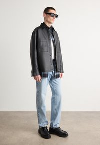 Chaqueta de cuero negra con dos bolsillos en el pecho, superpuesta a una camisa de cuadros, combinada con jeans de mezclilla azul claro y botas negras. Las gafas de sol completan el look.
