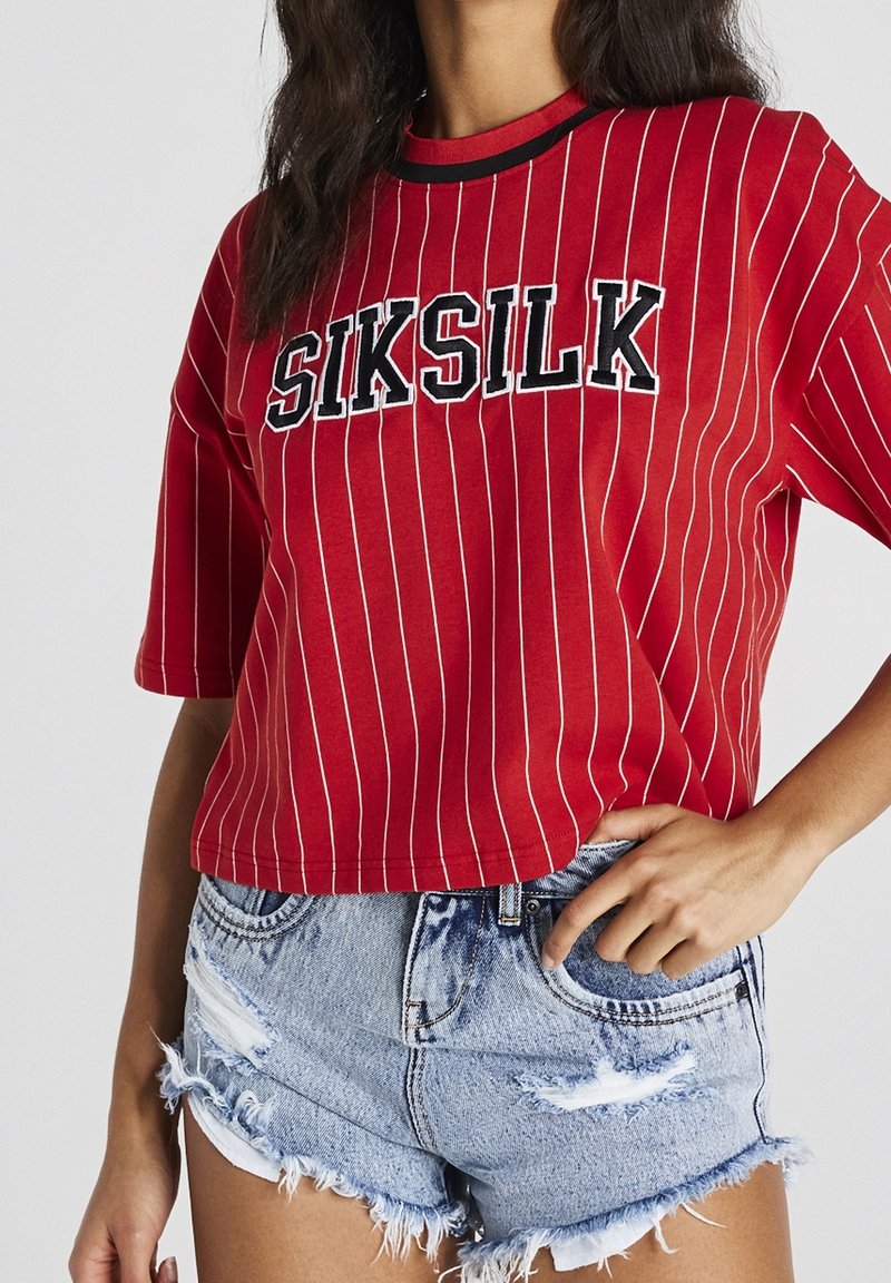 Punainen lyhythihainen t-paita, jossa on valkoisia pystyraitoja, ja jossa lukee sana "SIKSILK" näyttävillä mustilla kirjaimilla. Yhdistetty kulahtaneisiin farkkushortseihin.