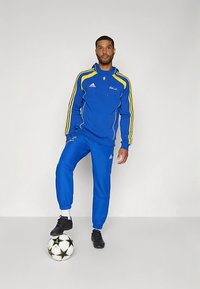 Trening albastru cu dungi galbene, hanorac cu fermoar și manșete elastice. Modelul stă cu un picior pe o minge de fotbal alb-negru.