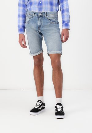Homme portant un short en denim bleu clair retroussé, une chemise à carreaux bleue, des chaussettes blanches et des baskets noires et blanches, debout sur un fond blanc.