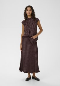 Top burdeos con lunares, cuello alto y mangas cortas combinado con una falda larga a juego. Tela suave con un diseño fluido; zapatos negros.