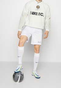 Biała koszulka Nike FC z długim rękawem i logo, białe spodenki, białe skarpetki oraz niebiesko-zielone buty piłkarskie na czarno-srebrnej piłce.