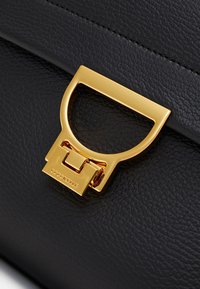 Sort læderhåndtaske med en markant guldfarvet metal-lås med geometrisk design og præget mærkelogo "Coccinelle."