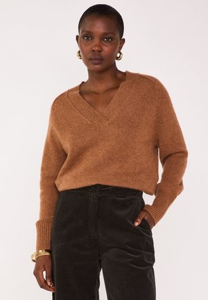 Vrouw met kort haar, die gouden ringoorbellen, een bruine V-halstrui, zwarte corduroy broek en gouden armbanden draagt, terwijl ze met één hand in haar zak staat.