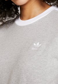 Grå bomullströja med en vit ribbad krage och en liten vit broderad logotyp på vänster sida. Enkelt, strukturerat tyg.