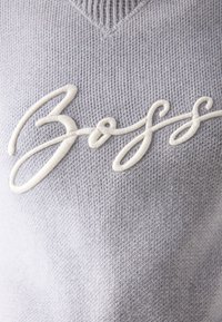 Tissu tricoté gris clair avec un texte en cursive blanche brodée en relief formant le mot « Boss » au centre.