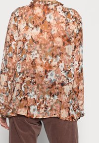 Blusa fluida con patrón abstracto multicolor que presenta tonalidades de marrón, blanco y azul. Detalle de escote con volantes y dobladillo tipo peplum.