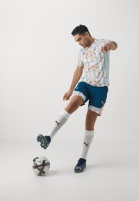 Jugador de fútbol vistiendo una camiseta con estampado y pantalones cortos azul oscuro, pateando un balón. La camiseta presenta detalles en naranja y un dorso azul claro.