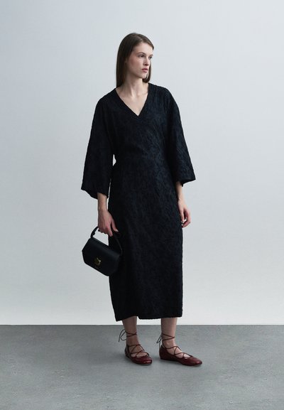 Robe noire avec un motif floral texturé, col en V et manches larges. Associée à des chaussures rouges à lacets et un petit sac à main noir.