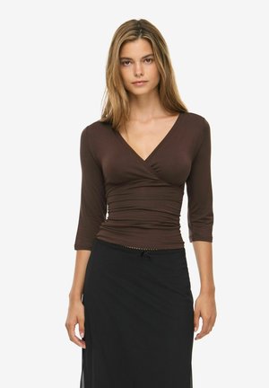 Femme aux cheveux blonds raides porte un haut cache-cœur marron à col en V avec des manches trois-quarts et une jupe noire sur fond blanc.