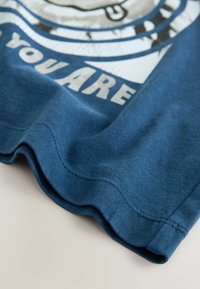 Sininen puuvillapaita, jossa on valkoinen graafinen printti. Kankaalla on sileä rakenne, jossa on kaareva helma ja näkyvät ompelusaumat.