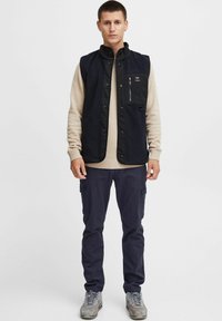 Chaleco de forro polar negro con cierres a presión y cremallera, que presenta un bolsillo en el pecho, usado sobre un suéter beige de canalé, combinado con pantalones oscuros y zapatillas grises.