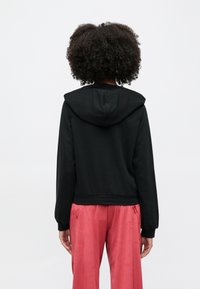 Göndör hajú személy hátulról, fekete kapucnis pulóvert és bő rózsaszín nadrágot visel, egyszínű világos háttér előtt.