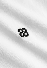 Petite forme de fleur abstraite noire brodée sur un fond de tissu blanc lisse avec texture de tissu visible.