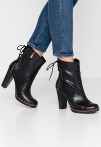 Stivali neri in pelle fino alla caviglia con punta affusolata, zip laterale e dettagli in pizzo, caratterizzati da un alto tacco a blocco e un particolare in legno.