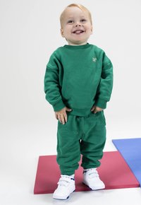 Groene sweatshirt en sweatsuit, gemaakt van zachte stof. Witte sneakers met blauwe accenten. Staande op kleurrijke matten, lachend kind.