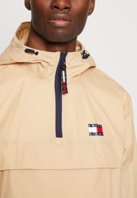 Lys beige vindjakke med halv lynlås, marineblå detaljer og et Tommy Jeans logo-patch. Stofudseendet virker glat og let.