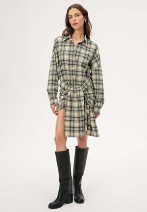 Kobieta ubrana w długą koszulę w kratę z węzłem z przodu oraz czarne kozaki za kolano, stojąca na jednolitym tle.