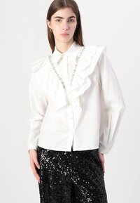 Camisa blanca con un delantero con volantes y detalles de joyas, combinada con pantalones de lentejuelas negros. Presenta mangas largas y cierre de botones.
