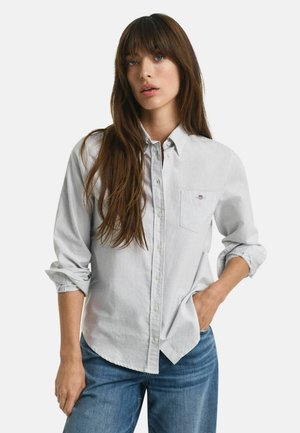 Frau mit langen braunen Haaren, die ein hell gestreiftes Button-Down-Hemd mit aufgerollten Ärmeln und blaue Jeans trägt, steht mit einer Hand in der Tasche.