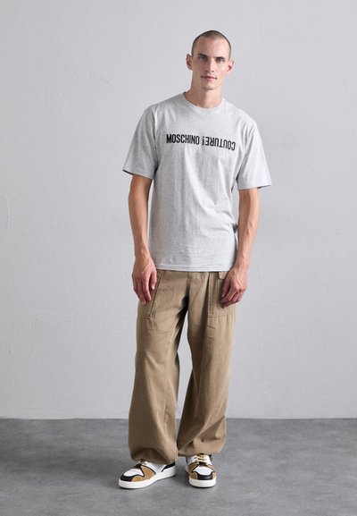 Tricou gri cu mânecă scurtă, cu inscripția "Moschino Couture!" imprimată în negru, asortat cu pantaloni cargo bej cu picioare largi și adidași albi cu accente.