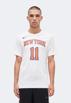 Jeune homme portant un t-shirt de sport blanc New York avec le numéro 11, debout devant un fond clair uni.