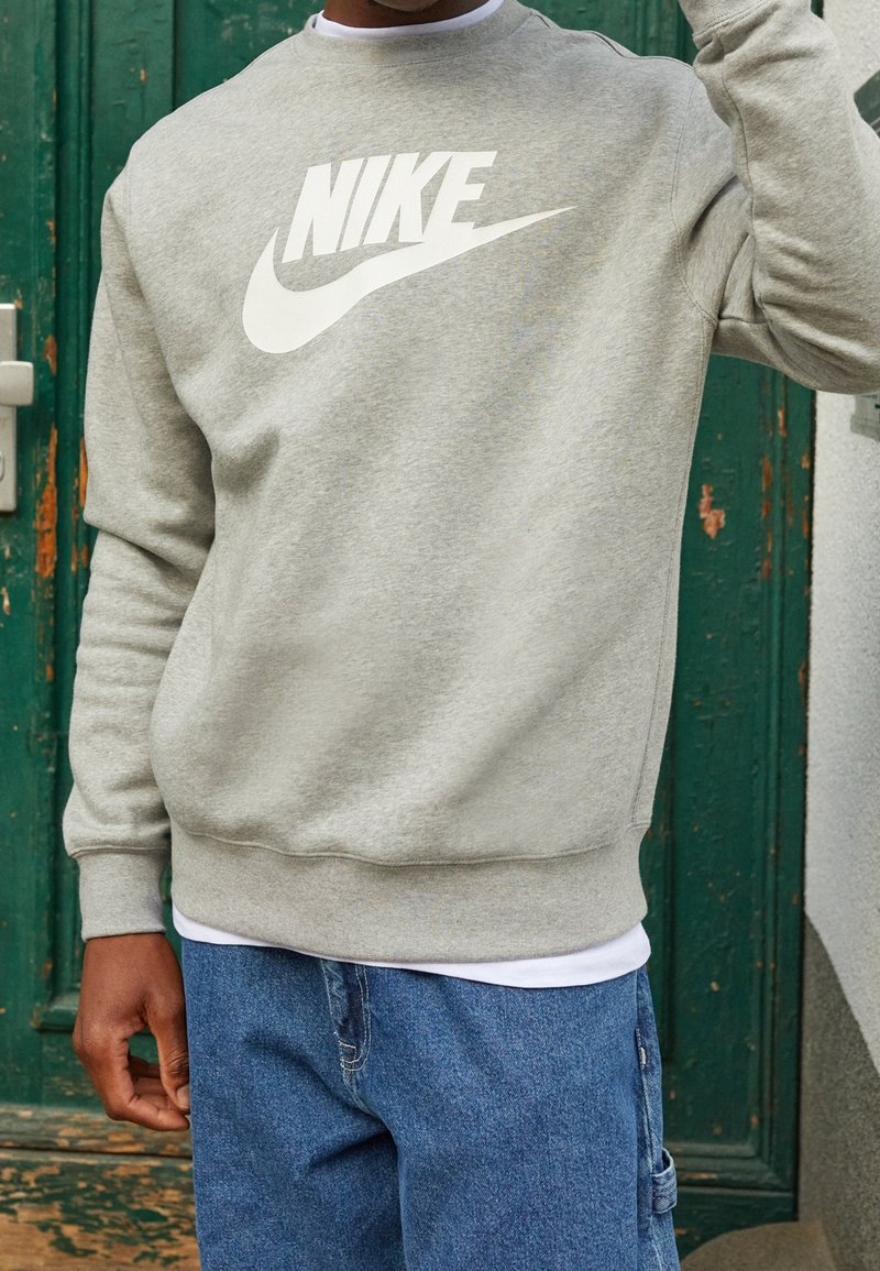 Grå sweatshirt med en stor vit Nike-logga, rund halsringning, ribbade muddar och fåll; bärs över en vit t-shirt med blå jeans.