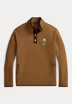 Jeune homme aux cheveux bouclés portant un pull marron, une chemise à carreaux, un jean bleu et des chaussures marron, debout les mains dans les poches.