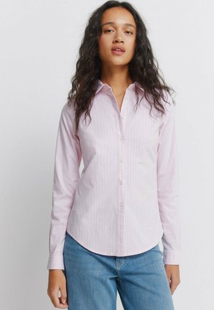 Pitkäaikaisiin laineisiin hiuksiin pukeutunut nainen, joka on vaaleanpunainen raidallinen nappipaita ja siniset farkut, seisoo yksinkertaisen valkoisen taustan edessä.