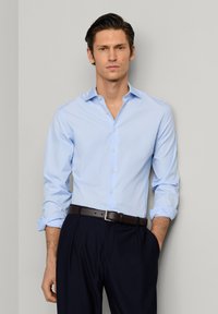 Junger Mann trägt ein hellblaues Hemd mit aufgekrempelten Ärmeln, schwarzen Gürtel und dunkle Hose, steht vor einer grauen Wand.