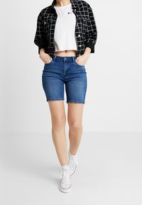 Modré džínové šortky s těsným střihem, v kombinaci s bílým zkráceným tričkem a černobílou kostkovanou košilí.