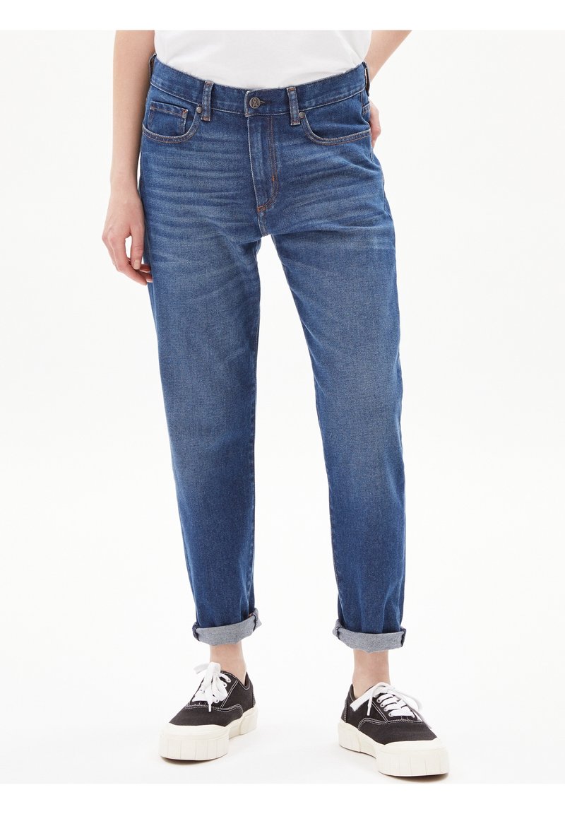 Person med uppvikta blå jeans och svarta låga sneakers med vita snören, som står mot en enkel vit bakgrund.