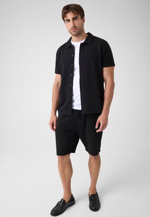 Mann trägt schwarzes kurzärmeliges Hemd mit Knöpfen über weißem T-Shirt, schwarze Shorts und schwarze Loafers, steht vor hellem Hintergrund.