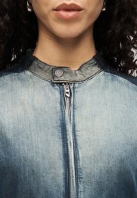 Τζιν μπουφάν με ασημένιο φερμουάρ, κλείσιμο με φερμουάρ μπροστά και όρθιο γιακά. Το ύφασμα έχει ξεθωριασμένη μπλε υφή με σκούρες πινελιές στον γιακά.