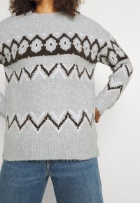 Pull en tricot gris clair avec un col rond, présenté avec un motif en zigzag marron et blanc sur la poitrine. Texture douce, coupe légèrement décontractée.
