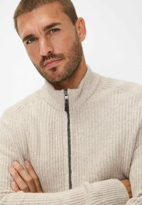Beige geripptes Strickpullover mit hohem Kragen und Reißverschluss vorne. Sanfte Textur und enganliegendes Design mit dunklem Reißverschluss.