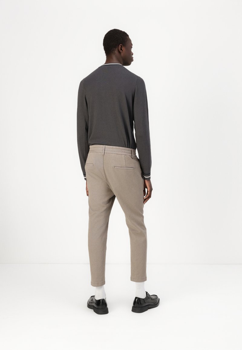 Graues langärmliges Hemd, beigefarbene maßgeschneiderte Hose, Seitentaschen, elastischer Bund, schwarze Schuhe und weiße Socken vor einem schlichten Hintergrund.