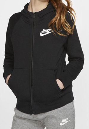 Person trägt einen schwarzen Nike Kapuzenpullover mit Reißverschluss und graue Nike Jogginghose, steht mit den Händen in den Taschen vor einem schlichten Hintergrund.