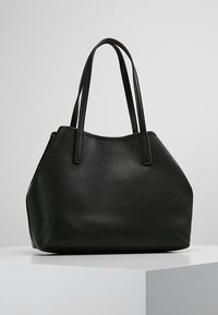 Sac en cuir noir texturé avec deux longues anses, présenté sur un piédestal blanc contre un fond gris uni.