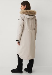 Cappotto invernale lungo beige con cappuccio foderato in pelliccia, vita elastica e dettagli neri ai polsini. Materiale liscio e resistente all'acqua con una vestibilità dritta.