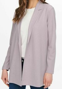 Blazer lavanda chiaro con colletto a punta, maniche lunghe e design aperto, realizzato in tessuto morbido, abbinato a una maglietta bianca.