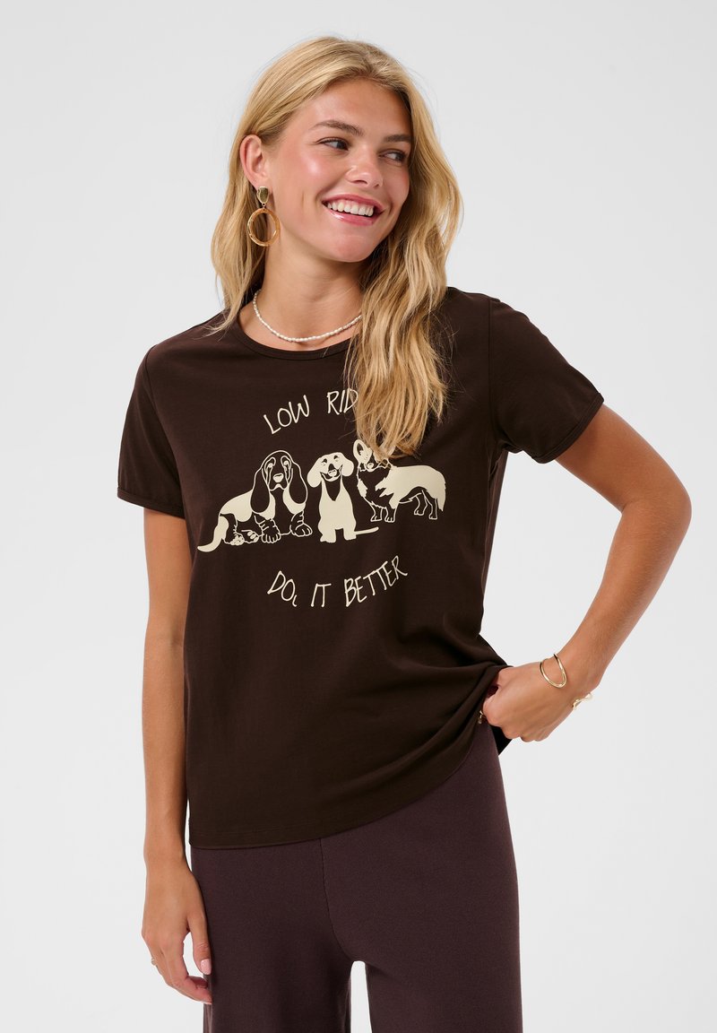 Jeune femme aux longs cheveux blonds portant un t-shirt marron foncé avec trois illustrations de chiens et le texte "Low Riders Do It Better."