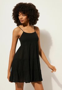 Schwarzes, ärmelloses Kleid mit tailliertem Oberteil und gestuftem, fließendem Rock. Aus leichtem Stoff mit strukturierter Oberfläche. Verstellbare Träger.